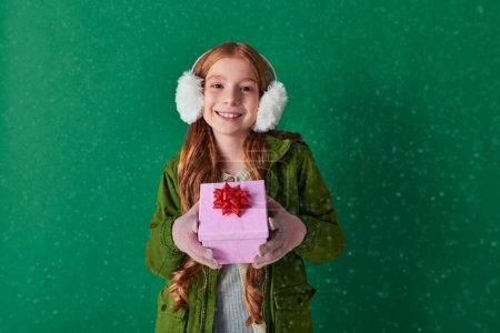 season of joy, happy kid in ear muffs and winter outfit holding Christmas gift under falling snow