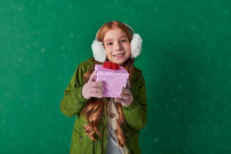 Jahreszeit der Freude, glückliches Kind in Ohrenschützern und Winteroutfit mit Weihnachtsgeschenk unter fallendem Schnee
