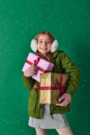 season of joy, happy girl in winter outfit and ear muffs holding holiday gifts under falling snow