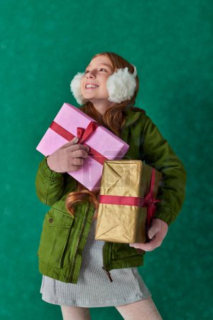 Téléchargez les photos : Saison de joie, fille heureuse en tenue d'hiver et cache-oreilles tenant des cadeaux de vacances sous la neige tombante - en image libre de droit