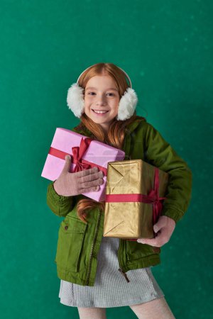 Freude, aufgeregtes Kind im Winteroutfit und Ohrenschützer mit Weihnachtsgeschenken unter fallendem Schnee