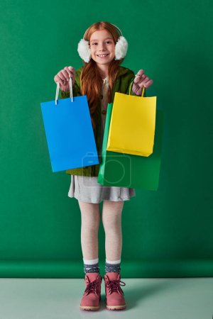 black friday and holiday season, cheerful kid in winter outfit and ear muffs holding shopping bags