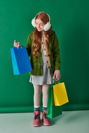 Foto de Viernes negro y temporada de vacaciones, niño feliz en traje de invierno y orejeras sosteniendo bolsas de compras - Imagen libre de derechos