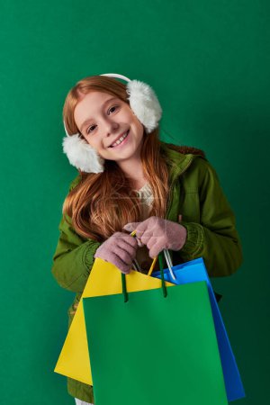 holiday season, joyful girl in winter outfit and ear muffs holding shopping bags on turquoise