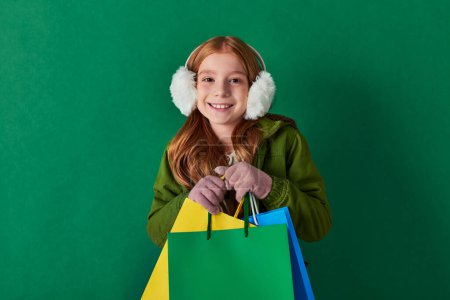 holiday season, excited kid in winter outfit and ear muffs holding shopping bags on turquoise