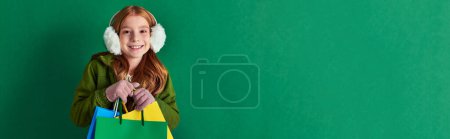Foto de Banner de temporada de vacaciones, niño feliz en traje de invierno y orejeras sosteniendo bolsas de compras en turquesa - Imagen libre de derechos