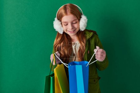 winter holiday, happy kid in winter outfit and ear muffs looking inside of shopping bag on turquoise
