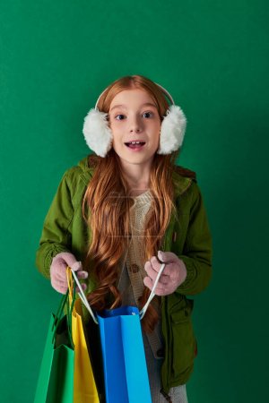 Winterurlaub, aufgeregtes Kind im Winteroutfit und Ohrenschützer mit Einkaufstaschen auf Türkis