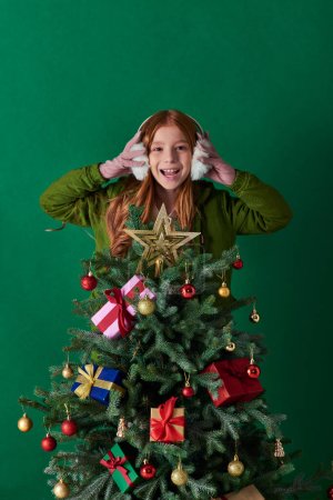holidays, excited girl wearing ear muffs and standing behind decorated Christmas tree on turquoise