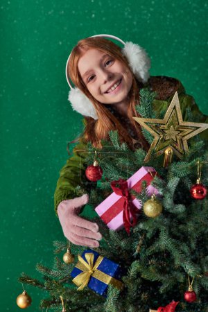 Feiertagsstimmung, fröhliches Mädchen im Ohrenschützer umarmt geschmückten Weihnachtsbaum vor türkisfarbener Kulisse