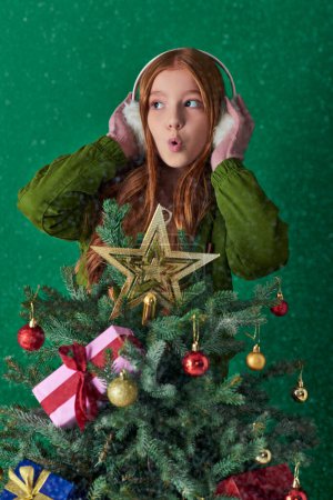 holiday spirit, surprised girl in ear muffs hugging decorated Christmas tree on turquoise backdrop