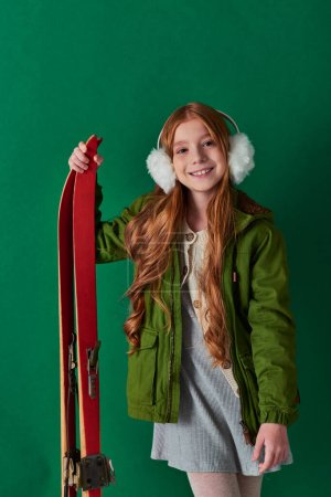 niña preadolescente feliz en orejeras y traje de invierno sonriendo y sosteniendo esquís rojos sobre fondo turquesa