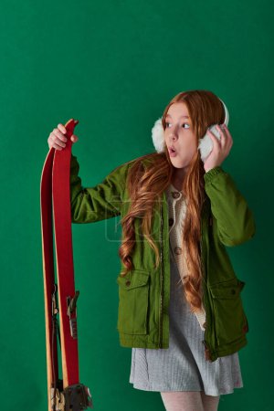 chica preadolescente sorprendido en orejeras y traje de invierno sosteniendo equipo de esquí rojo sobre fondo turquesa
