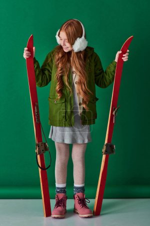 cheerful preteen girl in ear muffs and winter outfit holding red skis on turquoise background