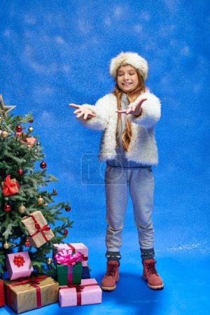 glückliches Mädchen in Kunstpelzjacke und Hut, das neben dem Weihnachtsbaum mit Geschenken auf blauem Schnee steht