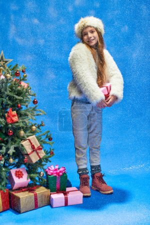 fröhliches Mädchen in Kunstpelzjacke und Hut mit Geschenk unter fallendem Schnee in der Nähe des Weihnachtsbaums auf blau