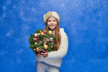 happy girl in faux fur jacket and hat holding decorated Christmas wreath under falling snow on blue