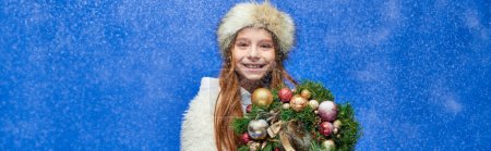 happy girl in faux fur jacket and hat holding decorated Christmas wreath under falling snow, banner
