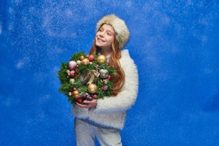 zufriedenes Mädchen in Kunstpelzjacke und Mütze mit Weihnachtskranz unter fallendem Schnee auf blau