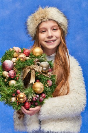 happy girl in faux fur hat and jacket holding Christmas wreath under falling snow on blue backdrop