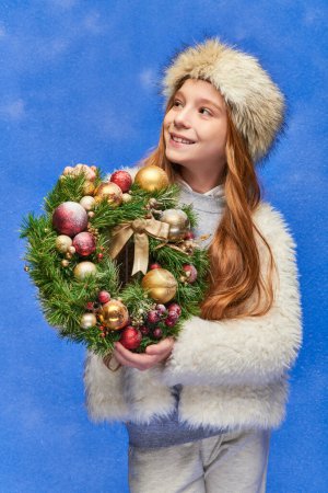 smiling girl in faux fur hat and jacket holding Christmas wreath under falling snow on blue