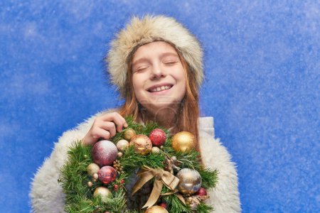 happy girl with closed eyes in faux fur hat and jacket holding Christmas wreath under falling snow