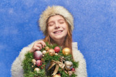 happy girl with closed eyes in faux fur hat and jacket holding Christmas wreath under falling snow Longsleeve T-shirt #680994062