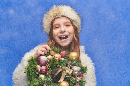 excited child in faux fur hat and jacket holding Christmas wreath under falling snow on blue