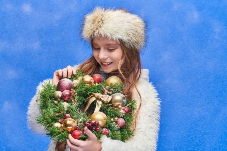 happy child in faux fur hat and jacket looking at Christmas wreath under falling snow on blue