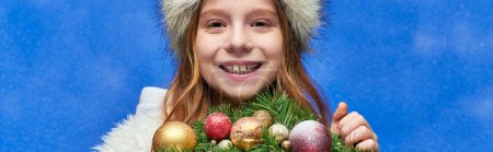 season of joy banner, happy girl holding Christmas wreath under falling snow on blue backdrop