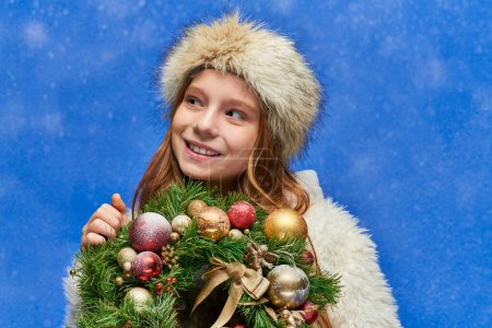 season of joy, cheerful preteen girl holding Christmas wreath under falling snow on blue backdrop