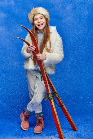 smiling preteen girl in winter faux fur jacket and hat holding red skis on turquoise background