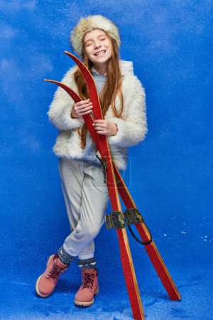 Photo for Winter joy, preteen girl in faux fur jacket and hat holding red skis on turquoise background - Royalty Free Image