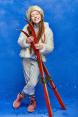 winter joy, preteen girl in faux fur jacket and hat holding red skis on turquoise background Mouse Pad 680994472