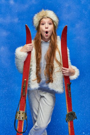 winter sport, shocked preteen girl in faux fur jacket and hat holding skis on turquoise background