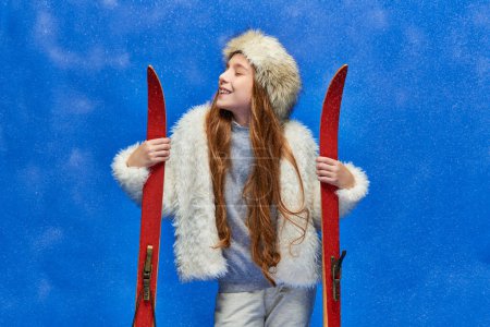 winter joy, pleased preteen girl in faux fur jacket and hat holding red skis on turquoise background