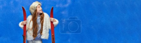winter joy, pleased preteen girl in faux fur jacket and hat holding red skis on turquoise, banner