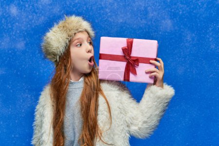 vacaciones de invierno, chica impactada en chaqueta de piel sintética y sombrero celebración caja de regalo sobre fondo turquesa