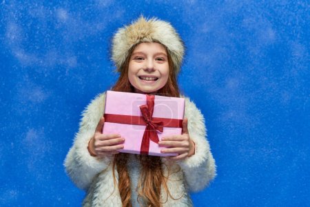 vacances d'hiver, fille heureuse en fausse veste de fourrure et chapeau tenant boîte cadeau enveloppé sur turquoise