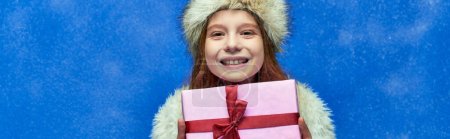 winter holiday banner, happy girl in faux fur jacket and hat holding wrapped gift box on turquoise