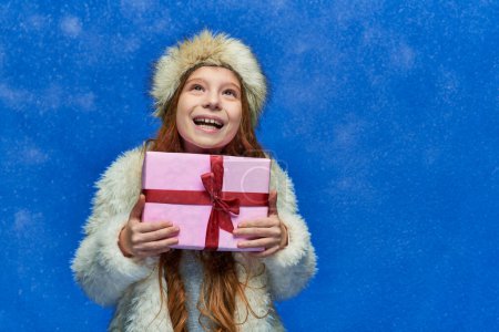 winter wonderland, happy girl in faux fur jacket and hat holding wrapped gift box on turquoise