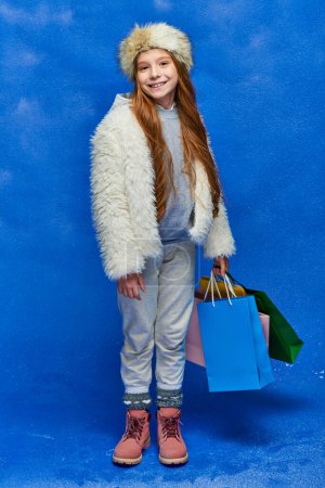 Foto de País de las maravillas de invierno, chica feliz en chaqueta de piel sintética y sombrero sosteniendo bolsas de compras en turquesa, nieve - Imagen libre de derechos