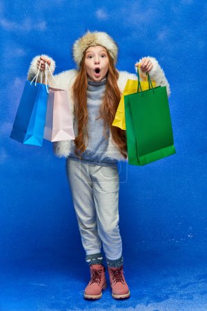 shopping de vacances, fille choquée en fausse veste de fourrure et chapeau tenant des sacs à provisions sur turquoise, neige