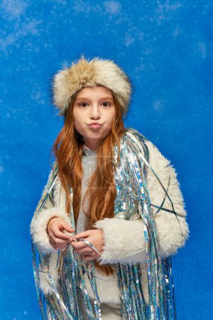 girl in faux fur jacket with tinsel standing under falling snow on blue backdrop, puffing cheeks