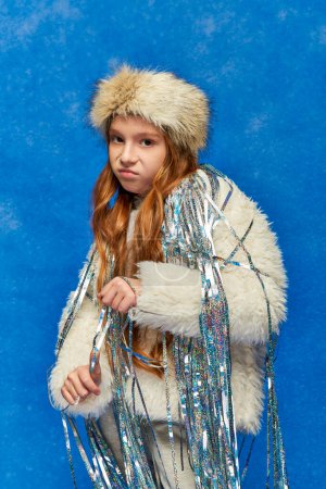 displeased girl in faux fur jacket with tinsel standing under falling snow on blue, feeling cold