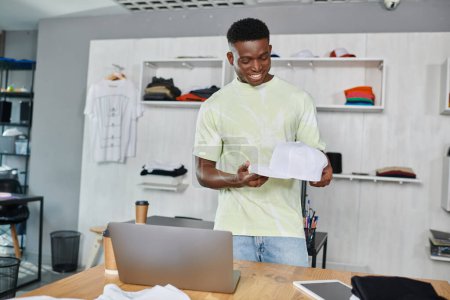 Foto de Sonriente diseñador afroamericano sosteniendo la tapa blanca cerca del ordenador portátil y la tableta digital en el estudio de impresión - Imagen libre de derechos