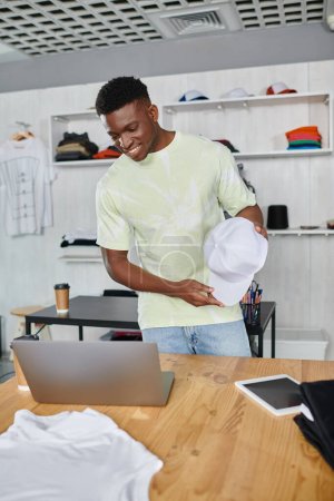 happy african american designer showing white cap during video call on laptop in print studio