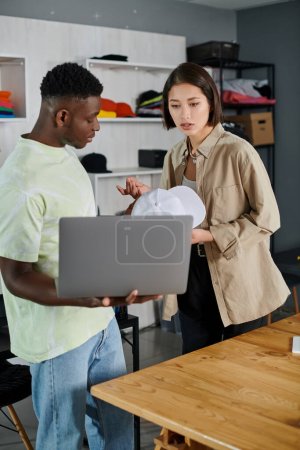 Foto de Jóvenes emprendedores interracial con gorra blanca y portátil discutiendo nueva startup en estudio de impresión - Imagen libre de derechos