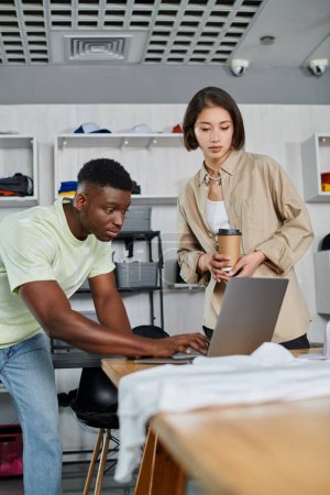 inspired african american man using laptop near asian woman with paper cup in clothing design studio