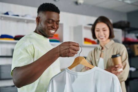 Foto de Alegre africano americano diseñador mostrando blanco camiseta sonriente asiático colega en impresión estudio - Imagen libre de derechos